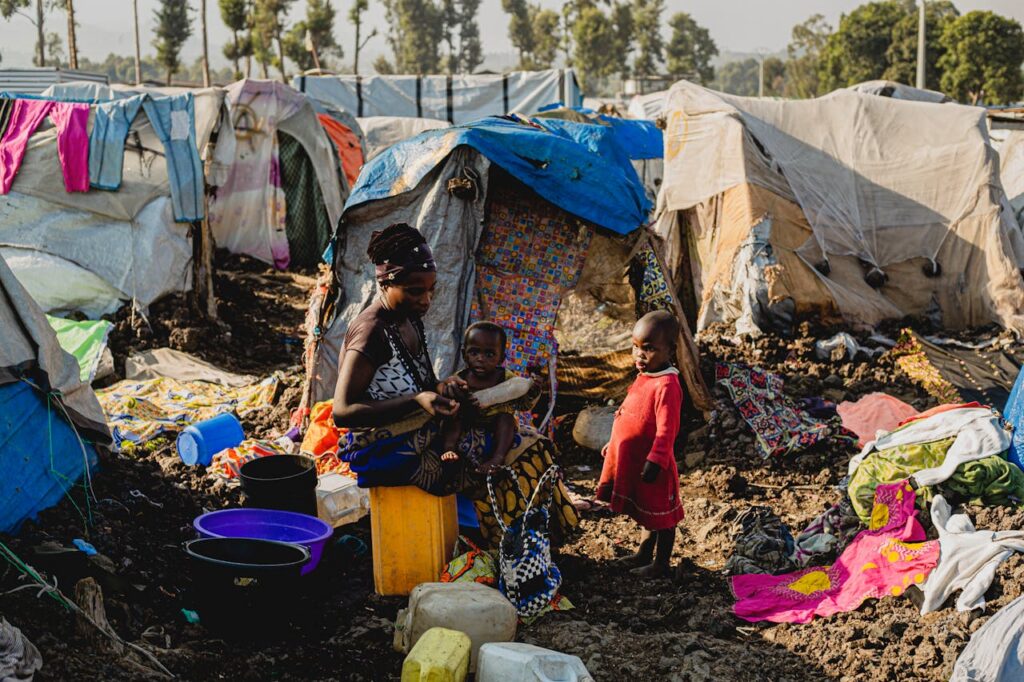Black woman with children in a makeshift camp setting amidst tents and scattered items.
